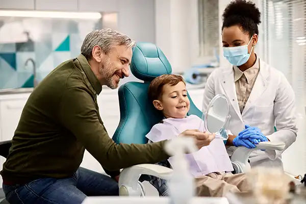 Parent holding a mirror for a happy, young child sitting in a dental chair while the dentist watches.