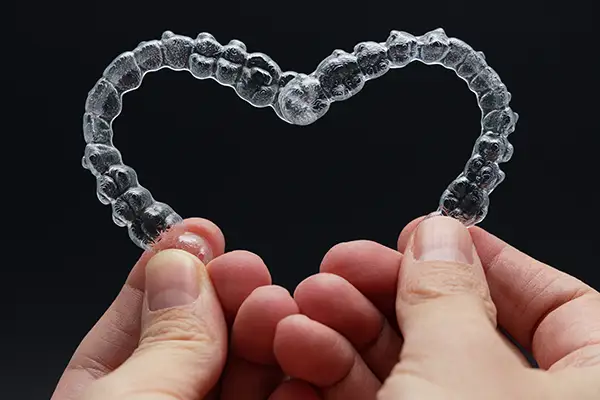 Hands forming a heart shape with two clear aligners against a black background, symbolizing love for straight, healthy teeth.