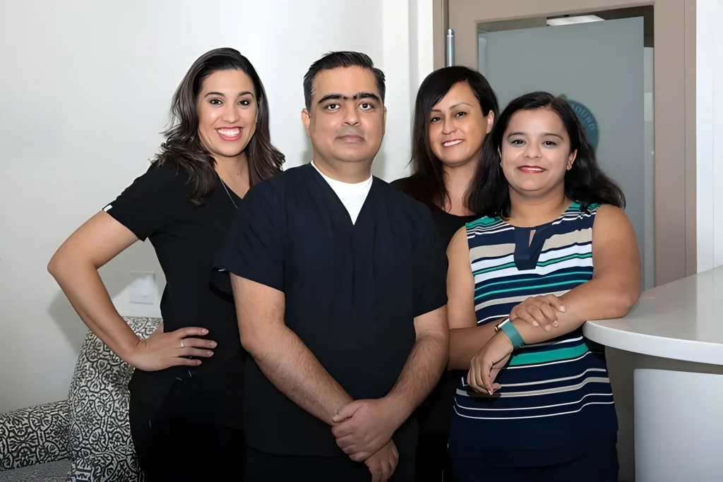 Four people stand together in a professional setting, three women and one man, all smiling or with neutral expressions, wearing business or medical attire, next to a counter and a door in an office environment.
