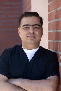 A man in dark scrubs and a white undershirt stands with arms crossed, leaning against a brick wall, looking at the camera with a neutral expression.