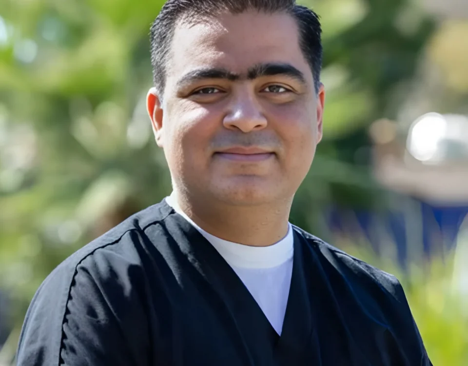 A man with short dark hair wearing black medical scrubs and a white undershirt stands outdoors, smiling slightly, with green foliage blurred in the background.