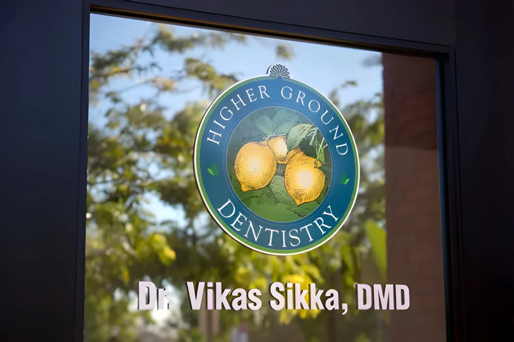 A glass door with the Higher Ground Dentistry logo featuring lemons, and the name Dr. Vikas Sikka, DMD, printed below it. Trees and sky are reflected in the glass.
