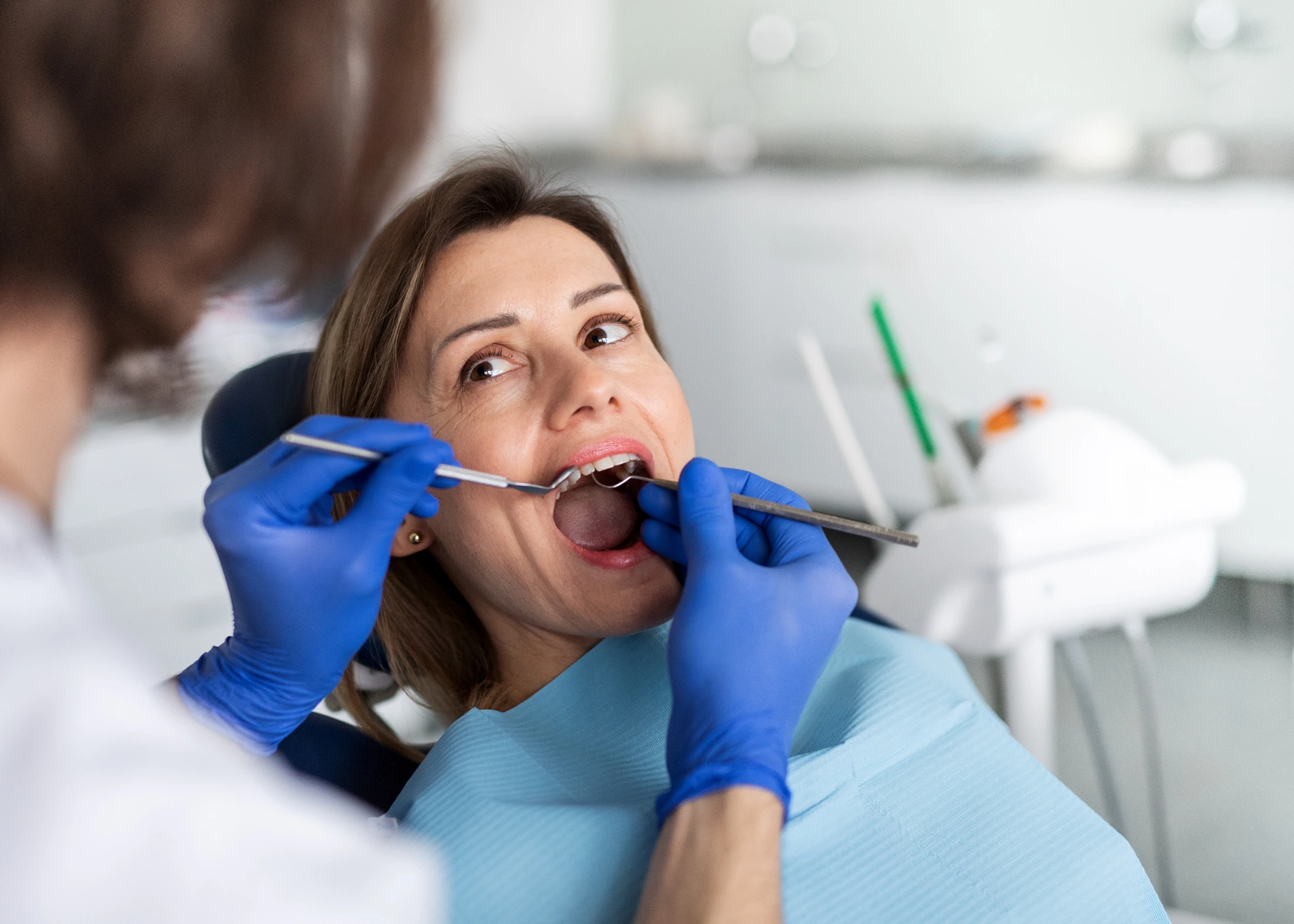 A woman sits in a dental chair with her mouth open while a dentist wearing blue gloves examines her teeth using dental tools.