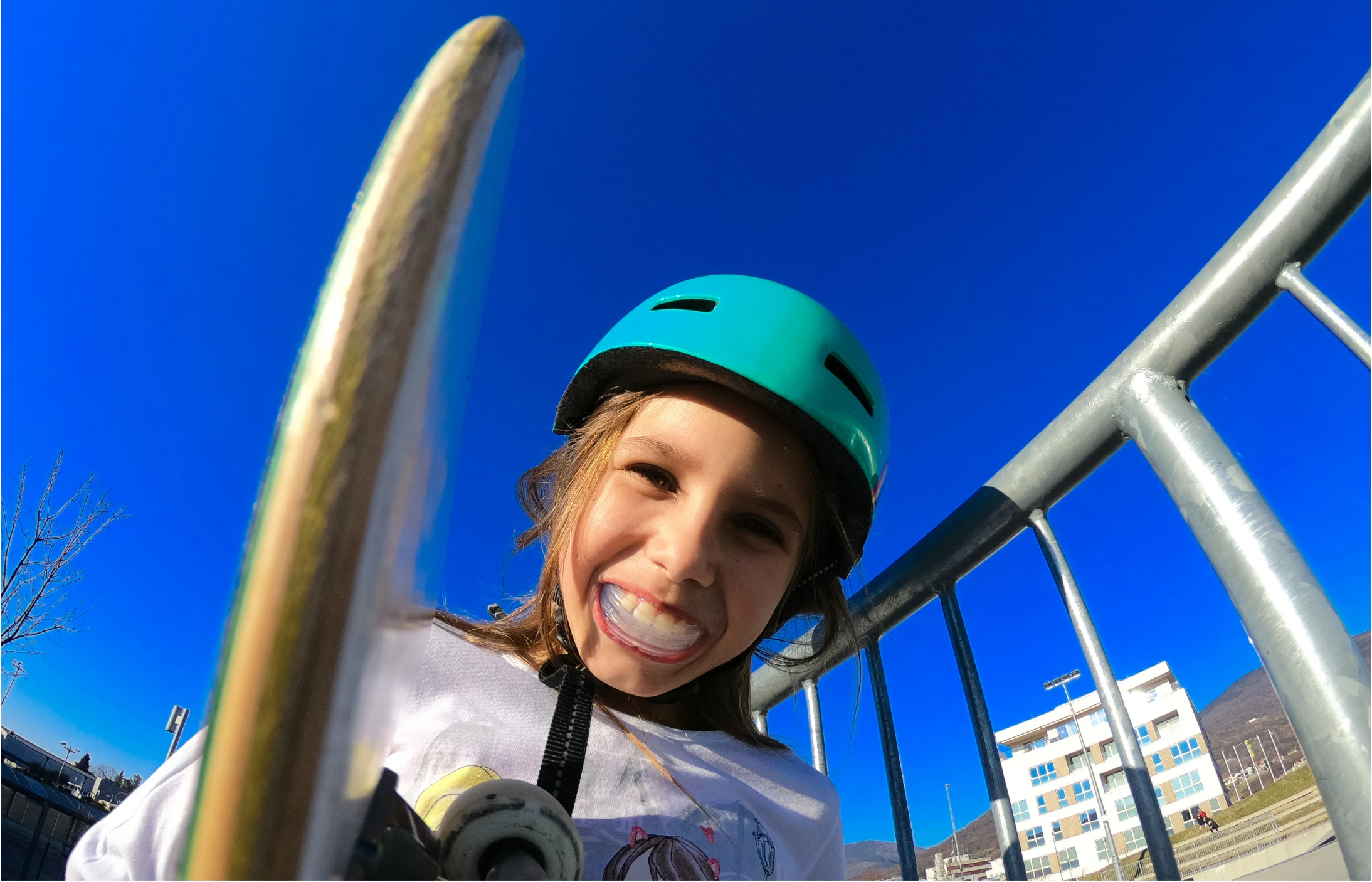 A young child wearing a teal helmet smiles widely while holding a skateboard at a skatepark, with a clear blue sky and metal railing in the background.