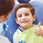 Smiling young boy in dental chair giving thumbs up with dentist in background