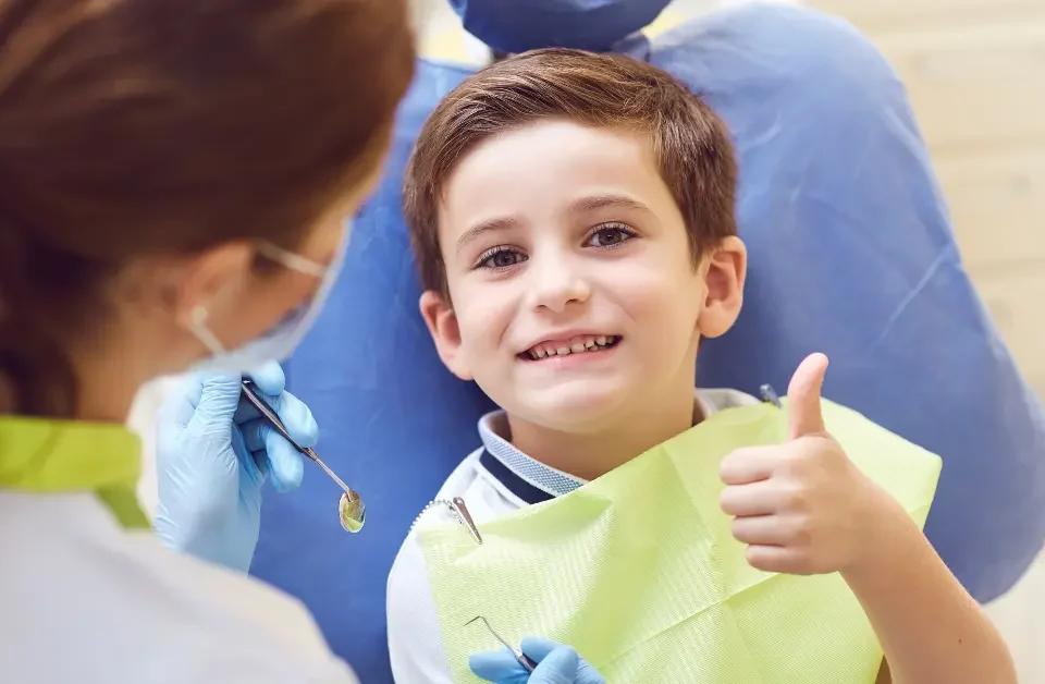 Smiling young boy in dental chair giving thumbs up with dentist in background