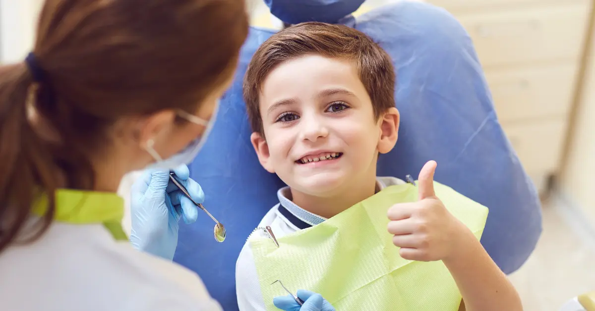 Smiling young boy in dental chair giving thumbs up with dentist in background