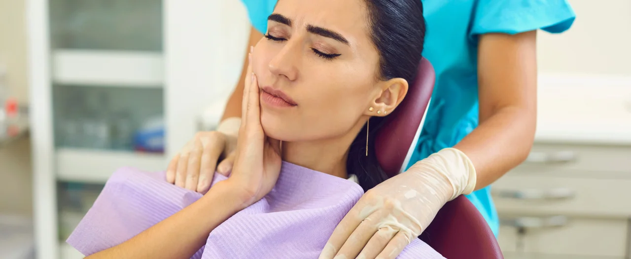 Woman in dental chair holding her jaw in pain with dentist standing beside her