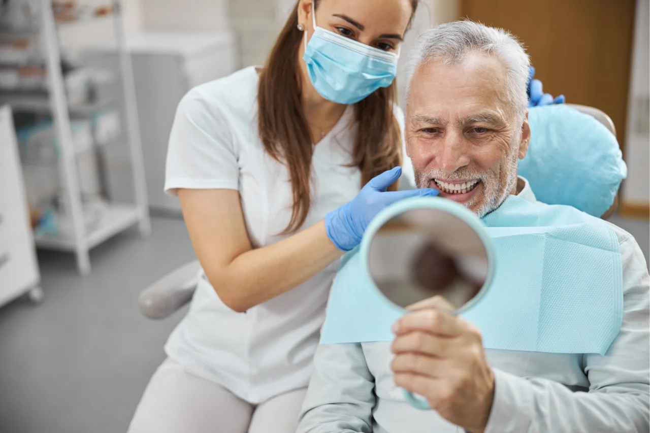 Senior patient in dental chair holding mirror and smiling while dentist stands beside him