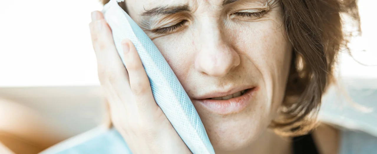 Woman holding ice pack against her cheek with eyes closed showing signs of tooth pain