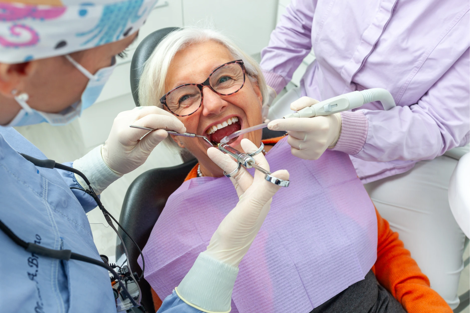 Senior woman wearing glasses in dental chair receiving oral examination from dentist and assistant