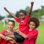 three smiling children in red soccer jerseys celebrate on a grassy field, with two kids lifting a third child who raises their arms triumphantly. in the blurry background, tmj upland emphasizes enamel protection for young athletes. a goalpost is visible behind them.