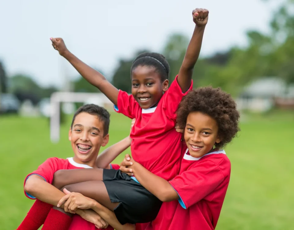 three smiling children in red soccer jerseys celebrate on a grassy field, with two kids lifting a third child who raises their arms triumphantly. in the blurry background, tmj upland emphasizes enamel protection for young athletes. a goalpost is visible behind them.