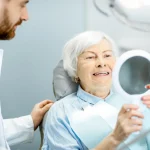 Senior woman smiling at mirror after dental implants procedure at Higher Ground Dentistry in Upland CA